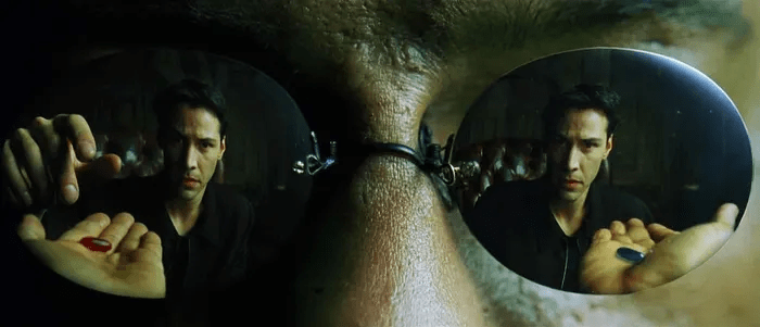 Still image from The Matrix showing Neo, reflected in Morpheus' glasses, reaching for the red pill in Morpheus' hand.